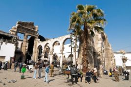 People sit around in a central square in Damascus. Ruins of ancient roman structures can be seen.